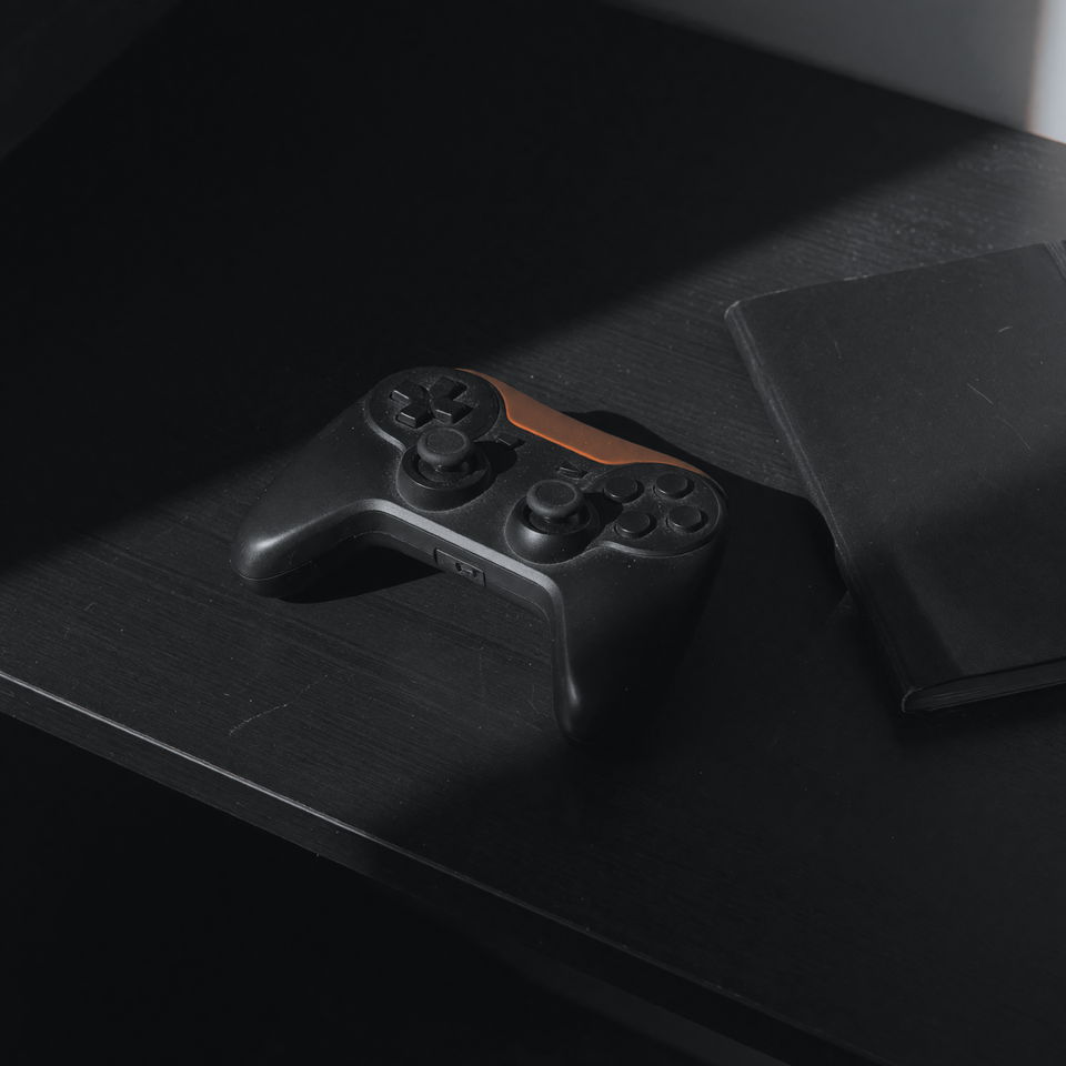 A matte black game controller on a dark desk beside a closed notebook, in hard low light.